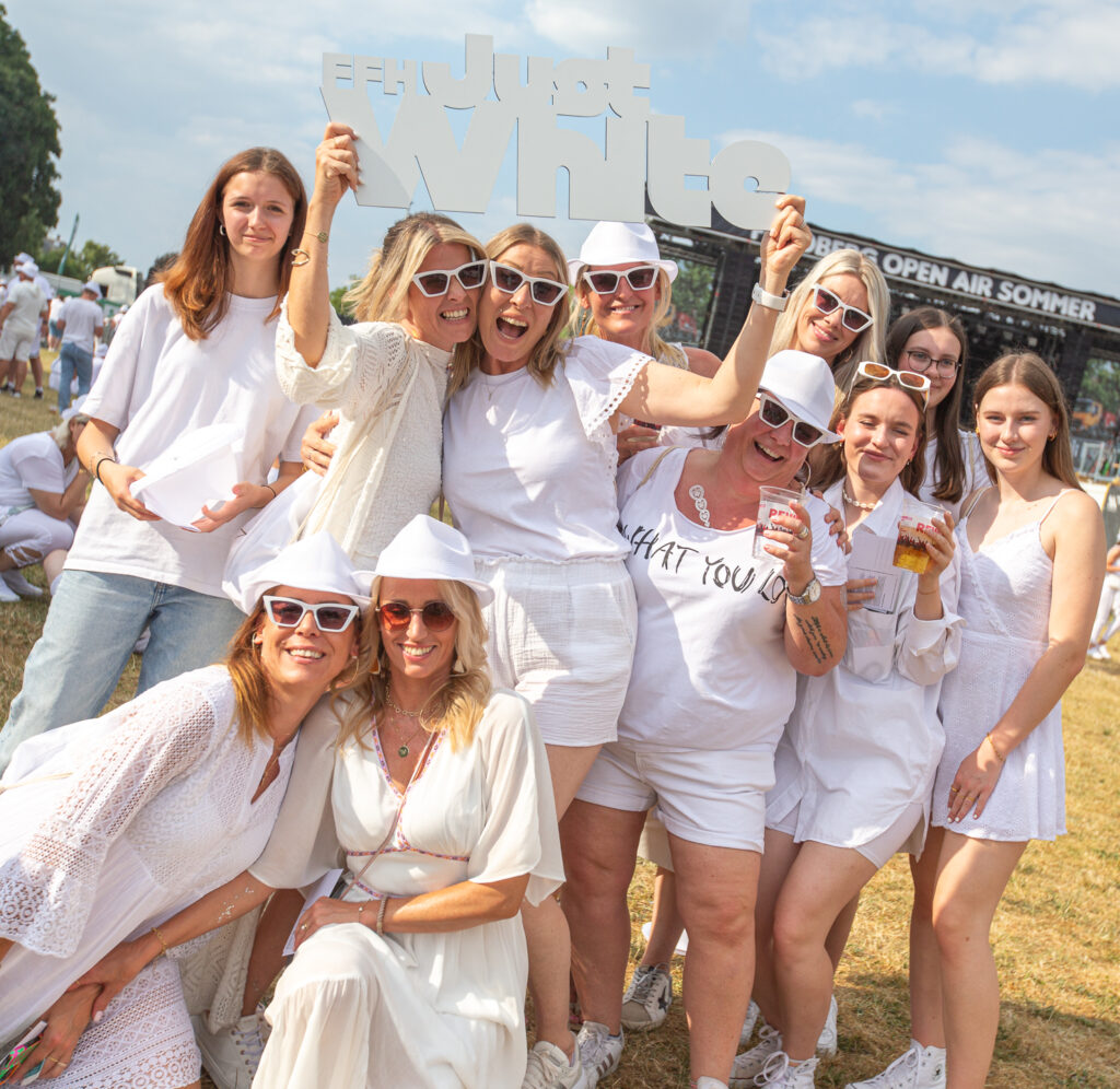 Eine Gruppe lächelnder, weiß gekleideter Frauen posiert auf einem Festival im Freien, einige tragen Sonnenbrillen und halten Getränke in der Hand, im Hintergrund sind eine Bühne und andere weiß gekleidete Menschen zu sehen.