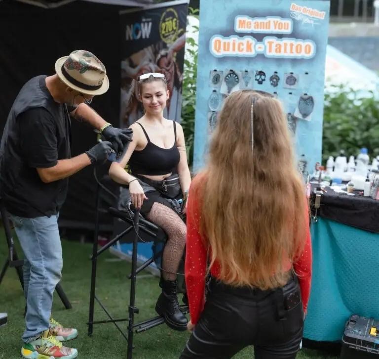 Ein Tätowierer bereitet sich darauf vor, einer sitzenden jungen Frau an einem Stand im Freien ein Tattoo zu stechen, während eine andere Frau mit langen Haaren in der Nähe steht und zusieht. Auf einem Schild steht "Me and You Quick Tattoo".