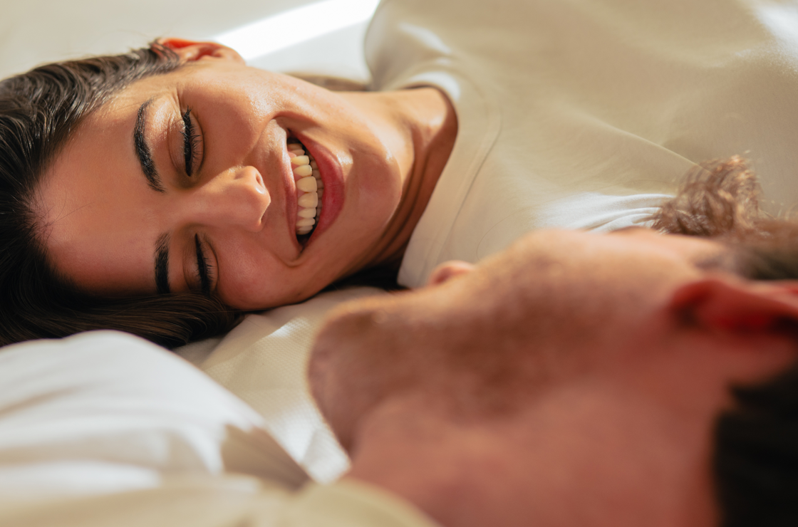 Eine Frau, die auf einem Bett liegt, l&auml;chelt eine andere Person neben sich warm an. Beide sind mit hellen Hemden bekleidet, und das sanfte Sonnenlicht beleuchtet ihre Gesichter.
