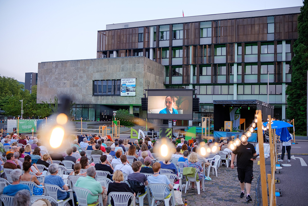 Eine Menschenmenge sitzt auf weißen Stühlen im Freien und sieht sich einen Film an, der auf eine große Leinwand vor einem modernen Gebäude projiziert wird, wobei Lichterketten im Vordergrund eine festliche Atmosphäre schaffen.