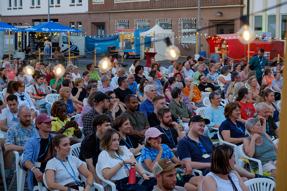 Eine große Gruppe von Menschen sitzt auf weißen Stühlen im Freien bei einer Veranstaltung und blickt aufmerksam nach vorne. Im Hintergrund sind Zelte, Essensstände und Lichterketten zu sehen. Die Atmosphäre wirkt lebendig und festlich.
