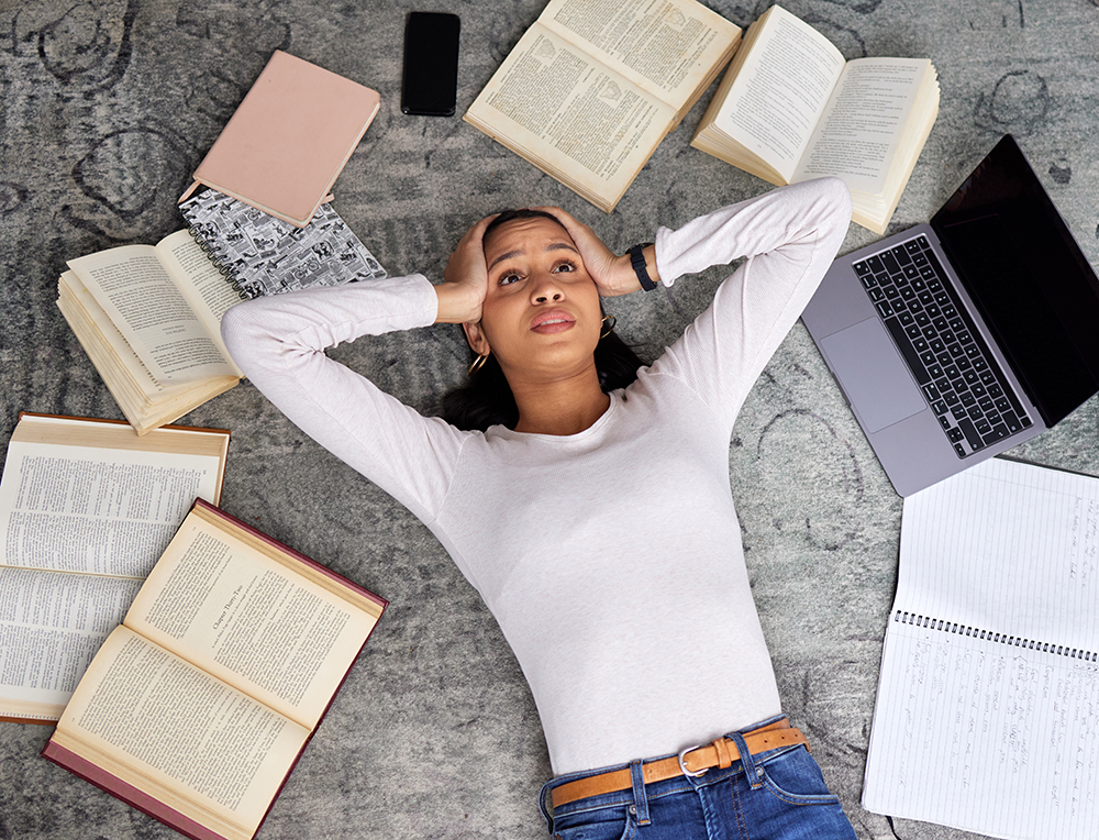 Eine Person liegt auf dem Boden, die Hände hinter dem Kopf, den Blick nach oben gerichtet, umgeben von aufgeschlagenen Büchern, einem Notizbuch mit Notizen, einem Laptop, einem Telefon und einem geschlossenen Notizbuch, was auf ein Studium oder ein Gefühl der Überforderung hindeutet.