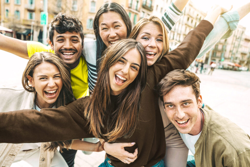 Eine Gruppe von sechs jungen Erwachsenen posiert eng beieinander im Freien, lächelnd und lachend, die Arme umeinander gelegt, und genießt einen sonnigen Tag in einer städtischen Umgebung.
