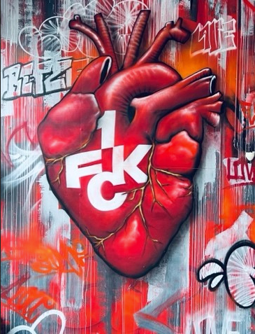 Ein Graffiti-Gemälde eines anatomisch detaillierten menschlichen Herzens an einer Wand, über dem in fetter weißer Schrift die Buchstaben 1 FKC stehen. Der Hintergrund ist mit bunter roter, weißer und schwarzer Sprühfarbe und Anhängern versehen.
