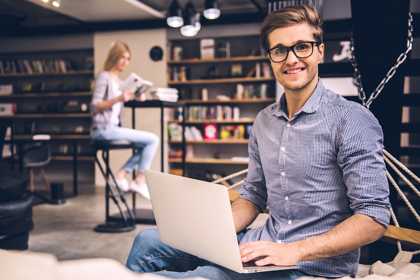 Ein junger Mann mit Brille sitzt lächelnd an einem Laptop in einer Bibliothek. Im Hintergrund liest eine Frau an einem hohen Tisch ein Buch. Hinter ihnen sind Bücherregale zu sehen.