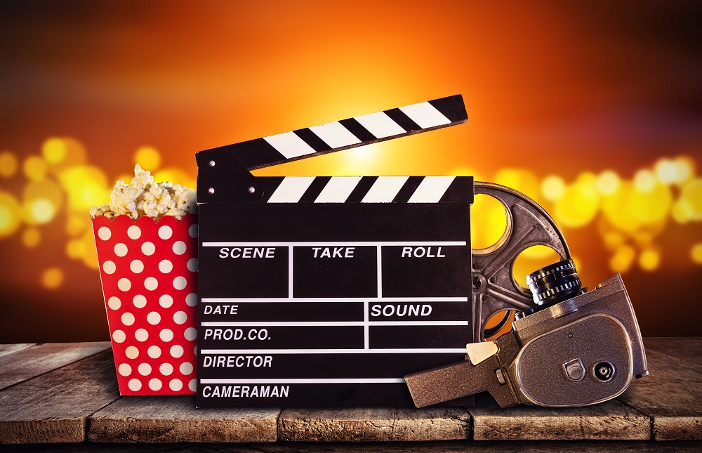 Eine Filmklappe, eine Vintage-Kamera, eine Filmrolle und eine rot-wei&szlig;e Popcornschachtel stehen auf einer Holzfl&auml;che vor einem leuchtenden, verschwommenen orangefarbenen Hintergrund.