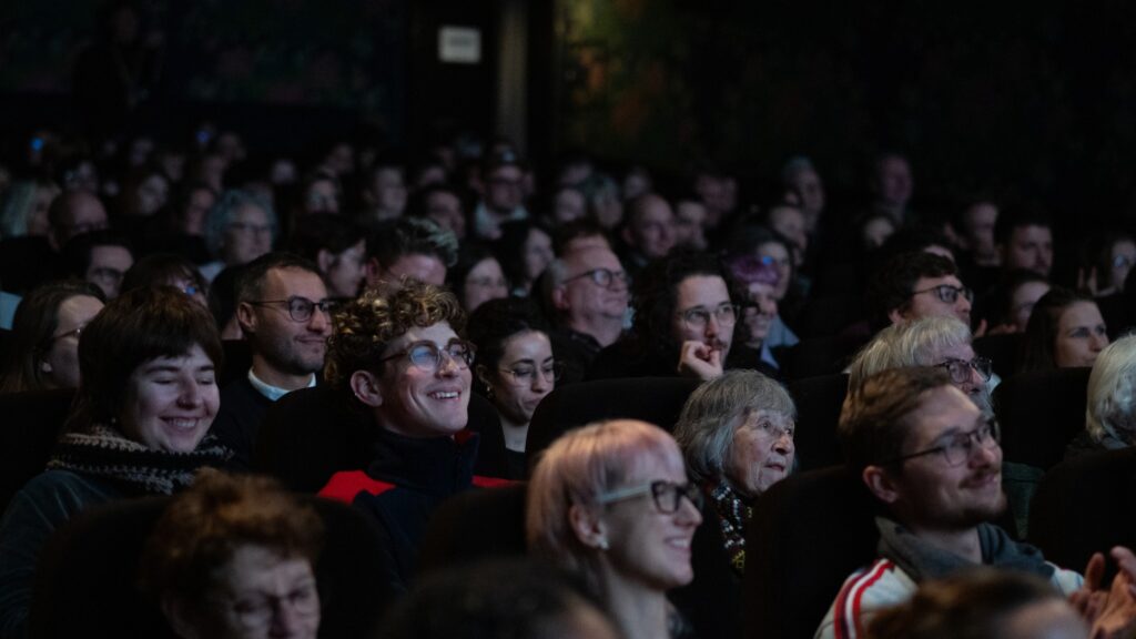 Ein bunt gemischtes Publikum unterschiedlichen Alters sitzt dicht beieinander in einem dunklen Kinosaal; viele lächeln und beobachten interessiert etwas außerhalb der Leinwand.