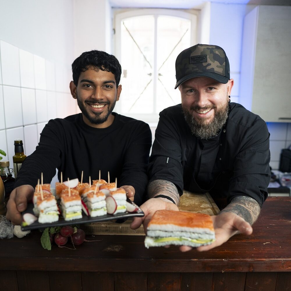 Zwei l&auml;chelnde M&auml;nner stehen hinter einer Holztheke in einer K&uuml;che; jeder h&auml;lt einen Teller mit ordentlich angeordneten Finger-Sandwiches oder H&auml;ppchen in der Hand und h&auml;lt ihn in Richtung Kamera.