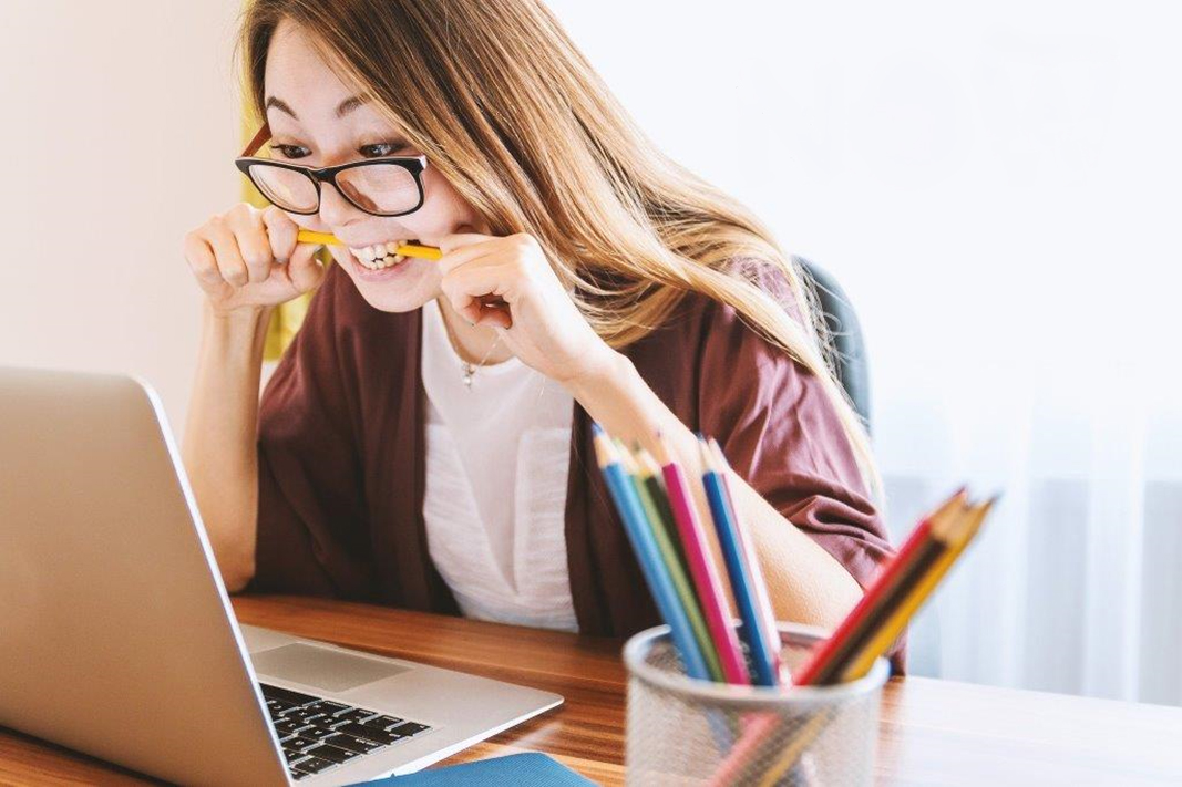Eine Frau mit Brille sitzt an einem Schreibtisch, beißt auf einen Bleistift und schaut dabei auf den Bildschirm eines Laptops. Auf dem Schreibtisch steht eine Dose mit bunten Bleistiften. Sie wirkt gestresst oder frustriert.