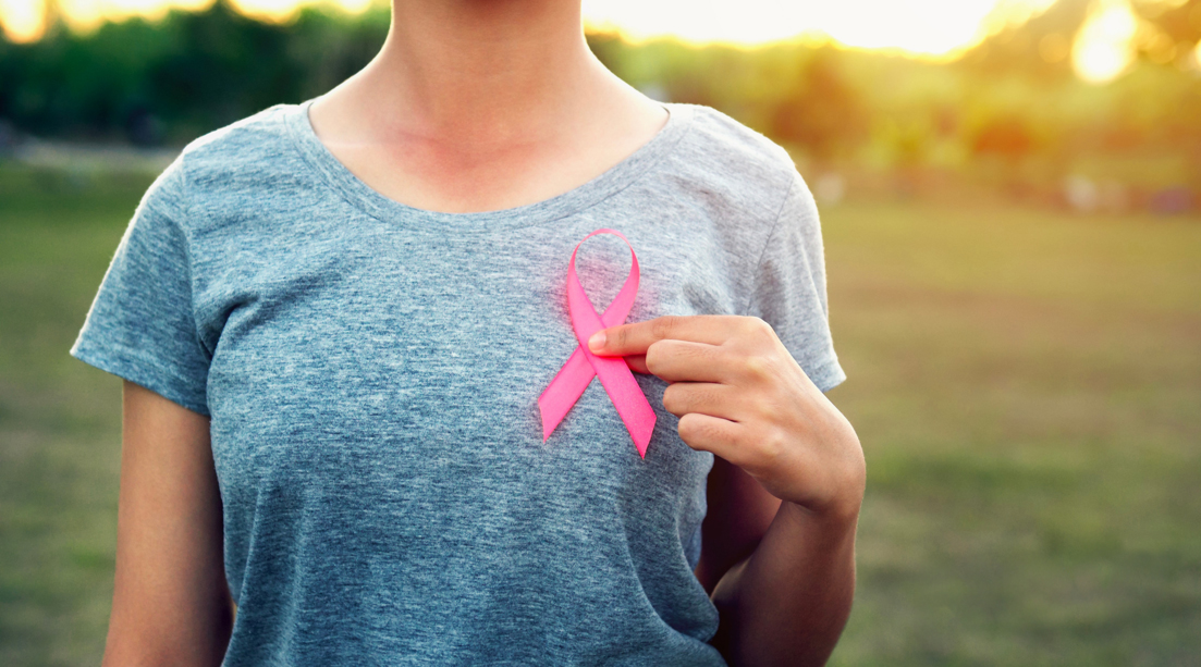 Eine Frau in einem grauen Hemd hält bei Sonnenuntergang im Freien eine rosa Schleife zur Sensibilisierung für Brustkrebs vor die Brust.
