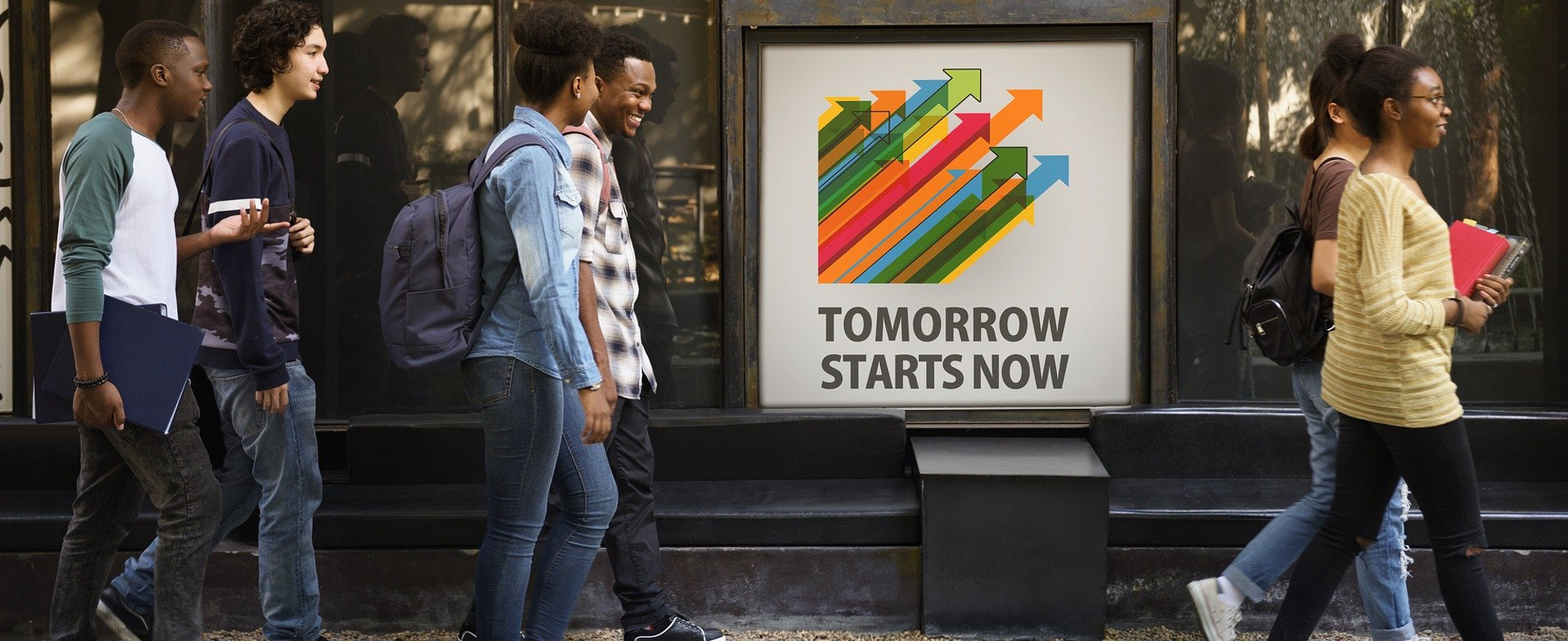 Eine Gruppe von Schülern geht an einem Gebäude vorbei. Hinter ihnen ist ein Plakat mit bunten, nach oben zeigenden Pfeilen und der Aufschrift „TOMORROW STARTS NOW“ zu sehen. Einige Schüler tragen Bücher und Rucksäcke.
