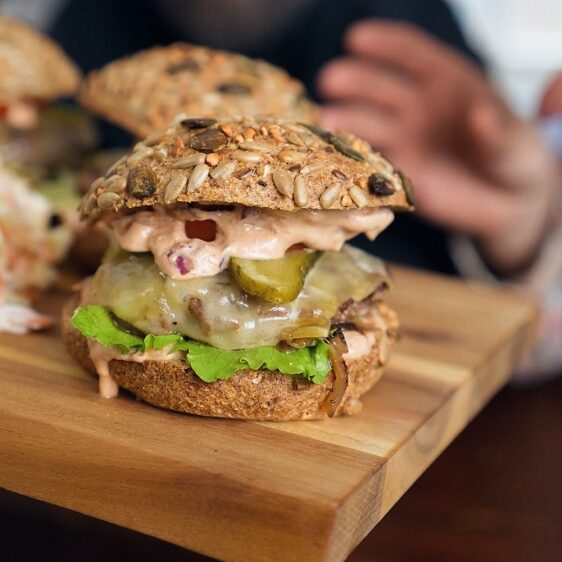 Eine Nahaufnahme eines Gourmet-Burgers mit Salat, Käse, Gewürzgurken und Soße auf einem Brötchen mit Körnern, serviert auf einem Holzbrett. Im Hintergrund streckt eine Person die Hand nach dem Burger aus und lächelt.