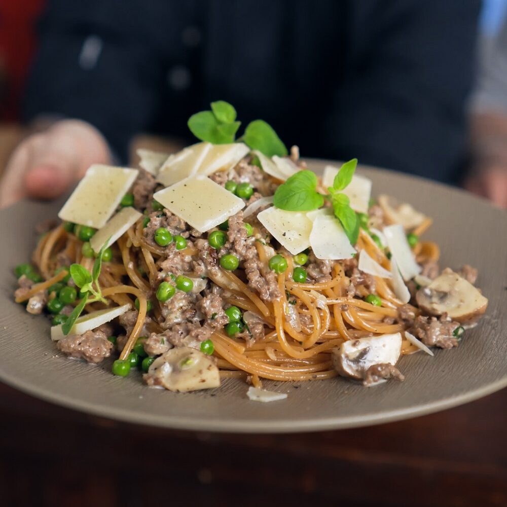 Eine Person, die einen Teller Spaghetti in der Hand hält, garniert mit Hackfleisch, Erbsen, Pilzen, frischem Basilikum und geriebenem Käse.