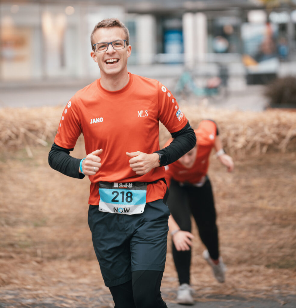 Ein lächelnder Läufer in einem roten T-Shirt mit dem Namen „Nils“ und der Startnummer 218 läuft im Freien, während ein weiterer Läufer hinter ihm herläuft und sich dabei nach vorne beugt. Beide befinden sich auf einem mit Stroh bedeckten Weg.