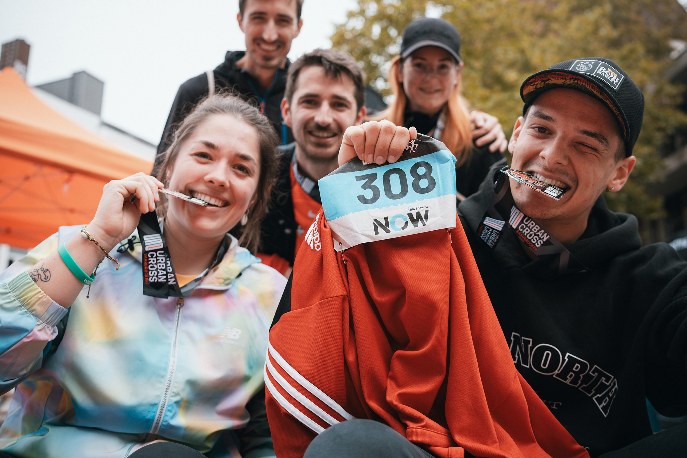 Fünf glückliche Menschen posieren nach einem Laufwettbewerb. Zwei von ihnen, die vorne sitzen, halten Medaillen, eine rote Jacke und eine Startnummer mit der Nummer 308. Die anderen lächeln im Hintergrund.